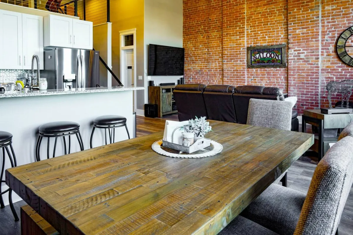 The photo features a cozy dining area with a large wooden dining table, brick wall backdrop, bar stools, a kitchen island, and a nearby television. The setup is warm and inviting, perfect for group meals, celebrations, or even working by the table.