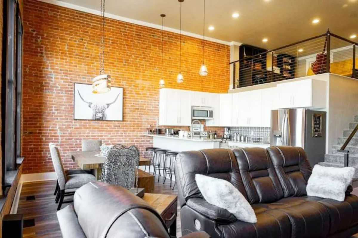 A view of an open-plan area where a modern kitchen flows into a cozy seating area, set off by an exposed brick wall and thoughtfully chosen artwork. The space feels both stylish and functional, perfect for hosting or relaxing.