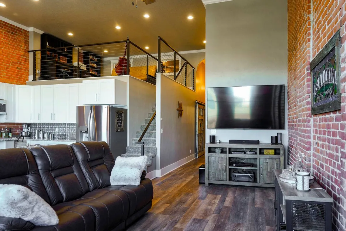 A spacious living area featuring a comfortable sofa, exposed brick wall, a modern television, and a stylish staircase. The room is bright and open, providing an ideal setting for relaxation or gathering with friends. The contemporary furnishings complement the historic architectural features.