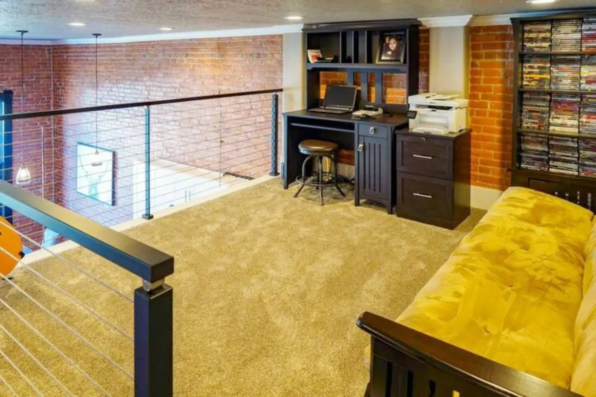The upstairs loft is depicted with a cozy workspace including a desk and computer, a comfortable yellow couch, an exposed brick wall, and a bookshelf stocked with DVDs. The space is inviting and creative, ideal for both working and relaxing.