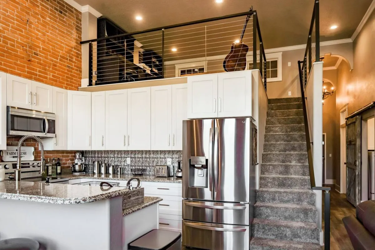 This image showcases a modern kitchen featuring a sleek granite countertop, stainless steel appliances, and an exposed brick wall. The open-plan design includes a staircase and is styled for contemporary comfort. The setting is perfect for both cooking and entertaining, with ample space for meal prep and socializing.