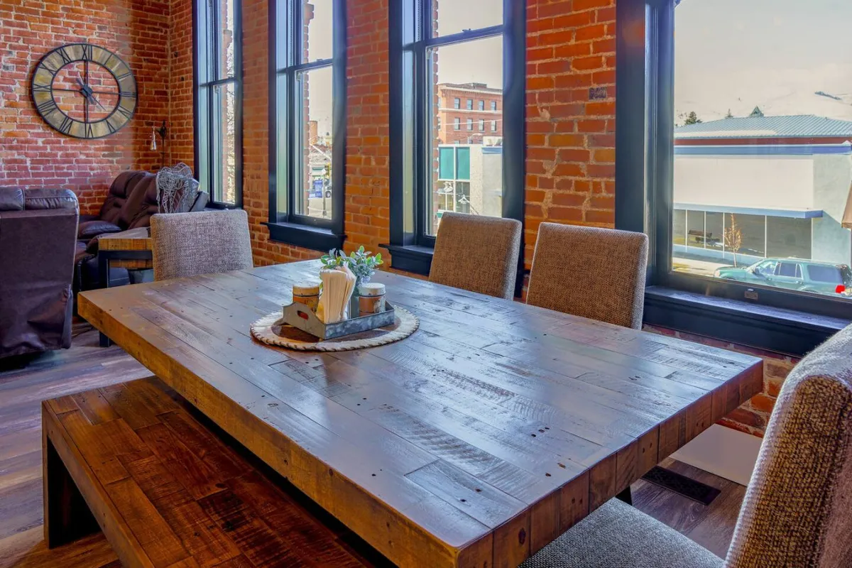 A cozy yet sophisticated dining space with a wooden table set against exposed brick walls, bathed in natural light from large windows. A classic clock adorns the wall, and the atmosphere feels inviting for socializing, dining, or relaxing while watching the activity on Main Street.