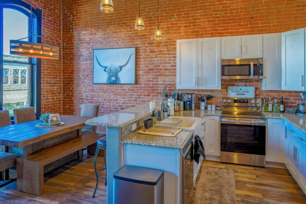 This inviting interior features a striking exposed brick wall, a warm wooden dining table perfect for gatherings, sleek modern kitchen appliances, and eye-catching artwork of a bull, all bathed in natural light to create a cozy yet contemporary ambiance.