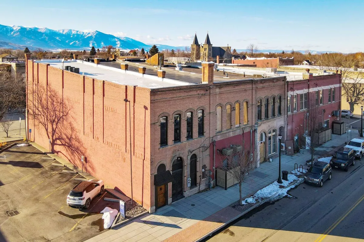 The historic brick facade of a downtown building with arched windows and a sidewalk shaded by trees, set against the scenic backdrop of surrounding mountains. The scene suggests architectural heritage, local history, and community engagement through events or outdoor activities.